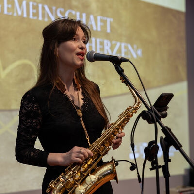 Bild vergr&ouml;&szlig;ern: Carmen Strobel steht in schwarzem Kleid mit einem goldfarbenen Saxofon am Mikrofon und singt. Im Hintergrund ist die goldene B&uuml;hnenprojektion mit dem Motto &raquo;Gemeinschaft im Herzen&laquo; zu sehen.