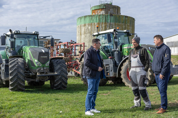 Bild vergr&ouml;&szlig;ern: Drei M&auml;nner stehen auf einer Wiese vor gr&uuml;nen Traktoren und landwirtschaftlichen Ger&auml;ten im Gespr&auml;ch. Der Mann in der Mitte tr&auml;gt eine graue Latzhose und eine M&uuml;tze. Dahinter steht ein hoher Betonsilo.