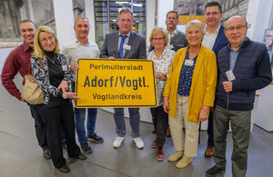 Bild vergr&ouml;&szlig;ern: Das Bild zeigt mehrere Akteure der Stadt Adorf. Sie halten ein gelbes Ortseingangsschild mit der Aufschrift "Perlmutterstadt Adorf/Vogtl. Vogtlandkreis" in die Kamera.