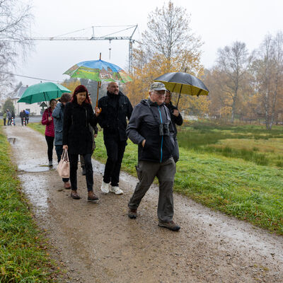 Bild vergr&ouml;&szlig;ern: Mehrere Besucher gehen mit Regenschirmen und wetterfester Kleidung &uuml;ber einen nassen Weg durch die herbstliche Landschaft bei Hammerbr&uuml;cke.