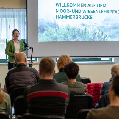 Bild vergr&ouml;&szlig;ern: Dr. Christine Heuck steht w&auml;hrend der Er&ouml;ffnungsveranstaltung vor den G&auml;sten und spricht an einem Mikrofon. Im Hintergrund ist eine Pr&auml;sentation zum Moor- und Wiesenlehrpfad Hammerbr&uuml;cke zu sehen.