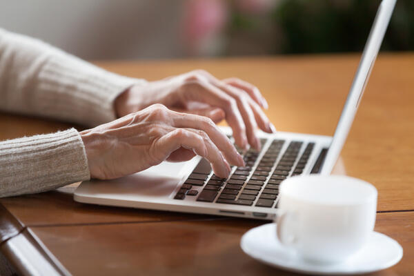 Bild vergr&ouml;&szlig;ern: Das Bild zeigt die H�nde einer �lteren Dame, die �ber der Tastatur eines Laptops schweben. Daneben steht eine wei�e Tasse auf einer Untertasse.