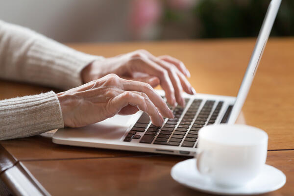 Bild vergrößern: Das Bild zeigt die Hnde einer lteren Dame, die ber der Tastatur eines Laptops schweben. Daneben steht eine weie Tasse auf einer Untertasse.