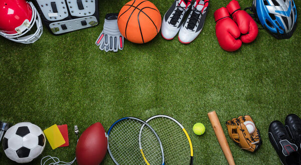 Das Bild zeigt eine Auswahl verschiedener Sportgegenstände auf einem grünen Rasen. Darunter sind ein Basketball, Federballschläger und Fußballschuhe. 