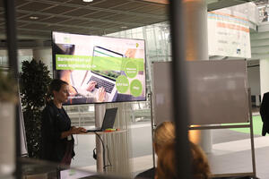 Bild vergrößern: Bianca Hecht stellte das Projekt Barrierefreiheit auf der landkreiseigenen Website www.vogtlandkreis.de vor. Sie steht hinter einem Laptop und im Hintergrund ist auf einem Monitor der Vortrag zu verfolgen.
